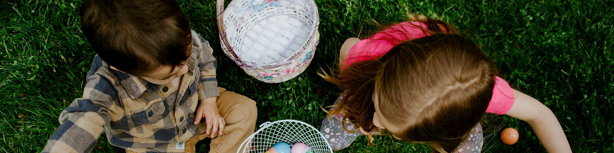 Two children sit on grass with baskets, collecting colorful Easter eggs scattered around them.
