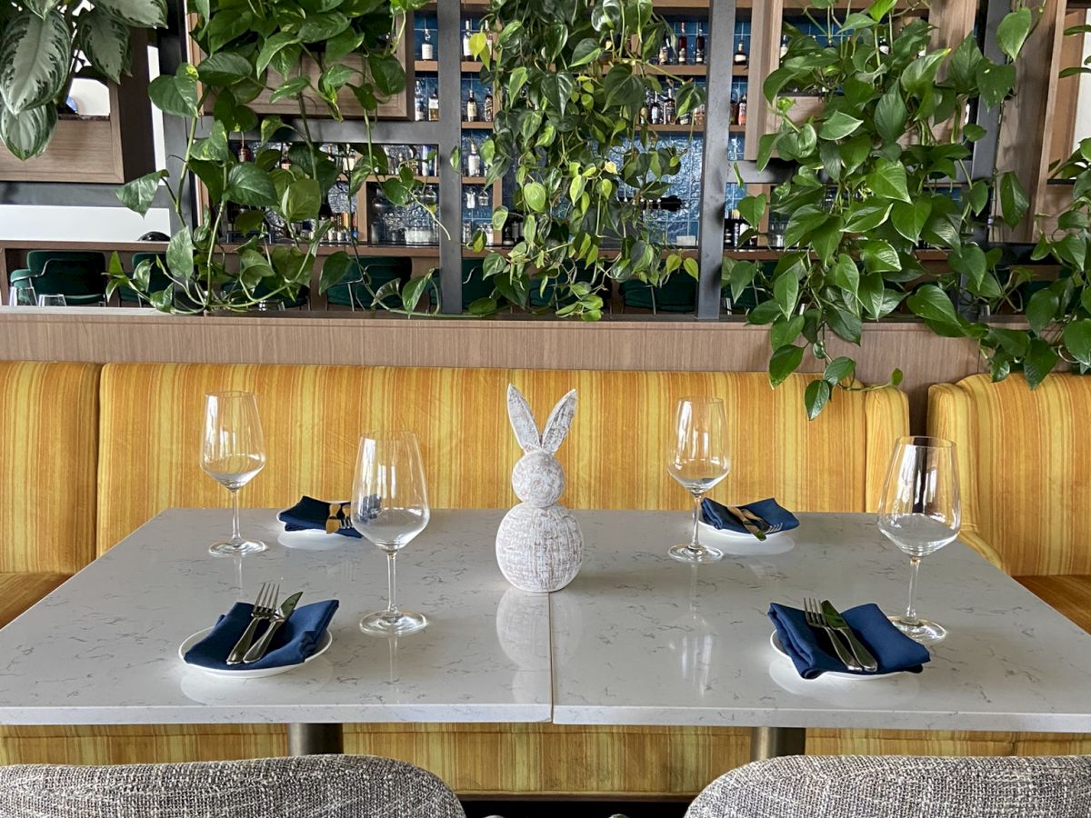 A table set for four with wine glasses and napkins, near a partition with hanging plants and a bar behind.