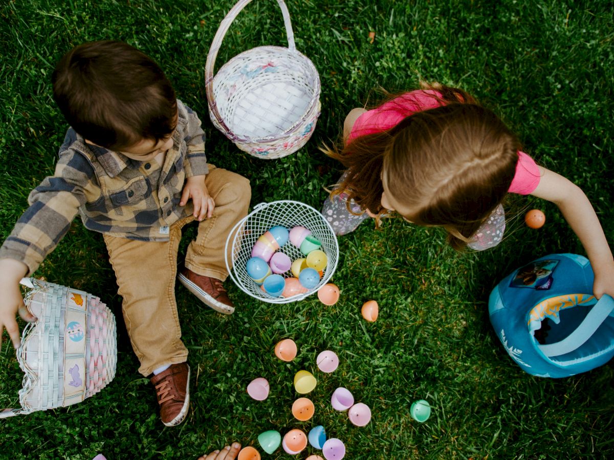 Two children are sitting on grass with baskets, collecting colorful Easter eggs scattered around them.
