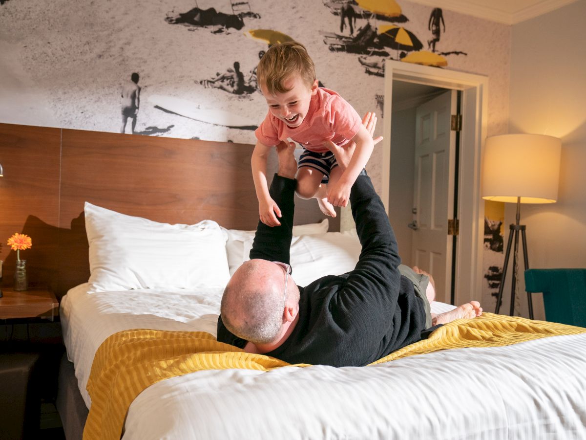 A person plays with a child, lifting them in the air while lying on a bed in a cozy room with a beach mural on the wall.