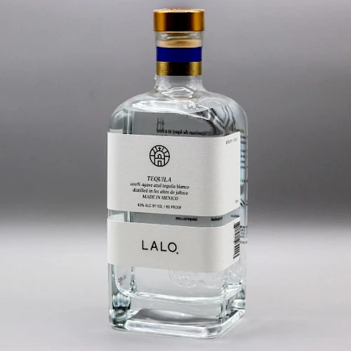 This image shows a bottle of LALO tequila, featuring a white label and a gold cap with a blue stripe, set against a neutral background.