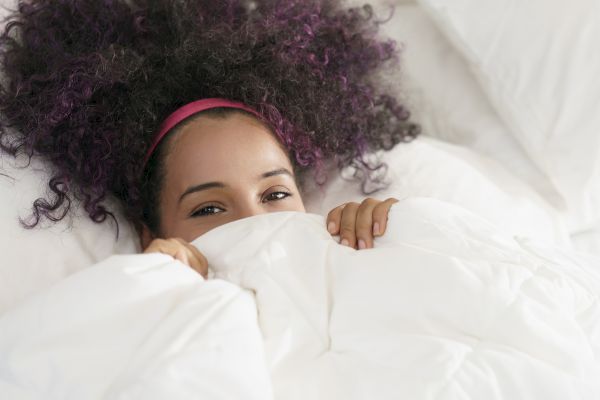 A person with curly hair and a pink headband peeks over a white blanket, lying in bed with a gentle expression.