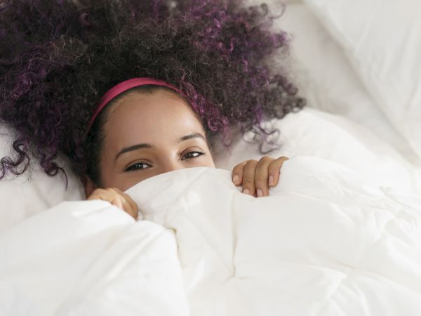A person with curly hair and a pink headband peeks over a white blanket, lying in bed with a gentle expression.