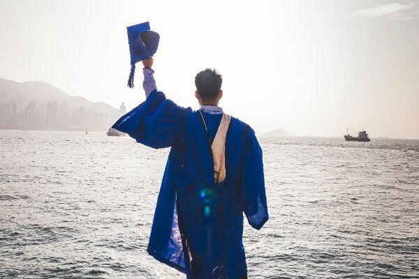 A person in a graduation gown, facing the water, holds a cap up triumphantly.
