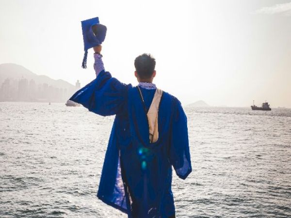 A person in a graduation gown, facing the water, holds a cap up triumphantly.