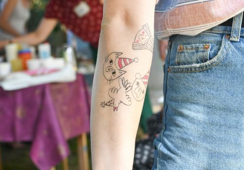 A person with cartoon-style temporary tattoos on their arm, wearing a striped shirt and jeans, standing near a table at an outdoor event.