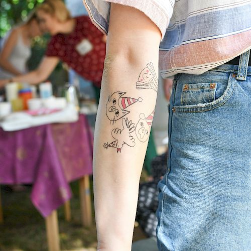 A person with cartoon-style temporary tattoos on their arm, wearing a striped shirt and jeans, standing near a table at an outdoor event.