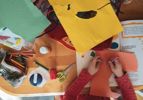 A child is crafting with colored paper, scissors, glue, and crayons on a table filled with art supplies and an open book.