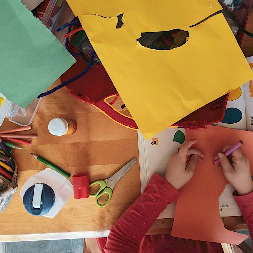 A child is crafting with colored paper, scissors, glue, and crayons on a table filled with art supplies and an open book.