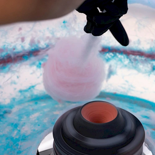A person is making pink cotton candy in a machine, with blue and pink sugar residue visible inside.