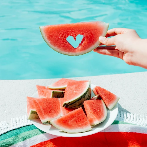 A hand holds a watermelon slice with a heart-shaped cutout, above a pool, next to a plate of watermelon slices on a towel.