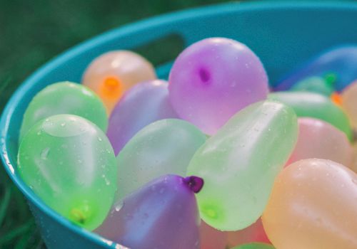 A blue bucket filled with colorful water balloons, some with droplets, sits on the grass.