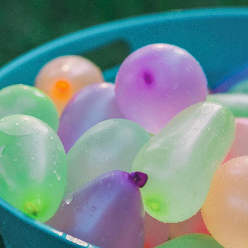 A blue bucket filled with colorful water balloons, some with droplets, sits on the grass.