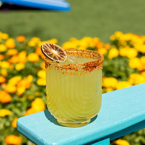 A yellow drink in a glass with a chili-rimmed edge and a dried lemon slice garnish sits on a teal surface, with flowers in the background.