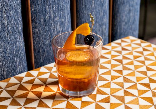 A cocktail in a glass garnished with an orange peel and a cherry on a skewer, placed on a geometric patterned table.