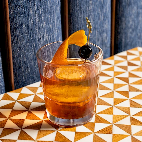 A cocktail in a glass garnished with an orange peel and a cherry on a skewer, placed on a geometric patterned table.
