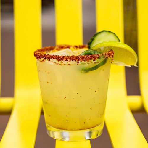 A yellow drink with a chili-rimmed glass, garnished with lime and cucumber, placed on a matching yellow chair for a vibrant look.
