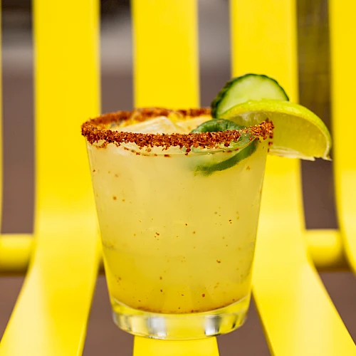 A yellow drink with a chili-rimmed glass, garnished with lime and cucumber, placed on a matching yellow chair for a vibrant look.