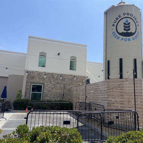 A building with a large silo labeled "Pure Project Beer For Good" is surrounded by wooden fencing and string lights.