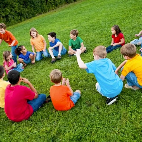 A group of children wearing colorful shirts sit in a circle on a grassy field, engaging in outdoor activities together.