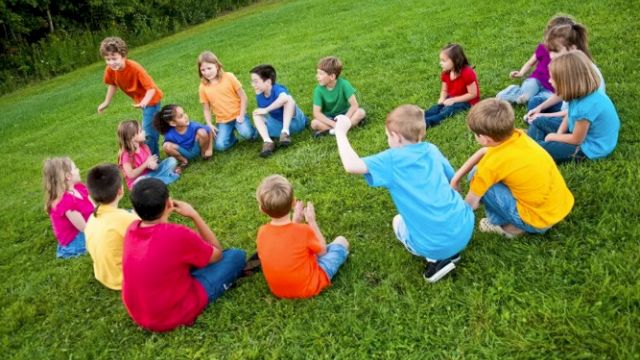 A group of children wearing colorful shirts sit in a circle on a grassy field, engaging in outdoor activities together.