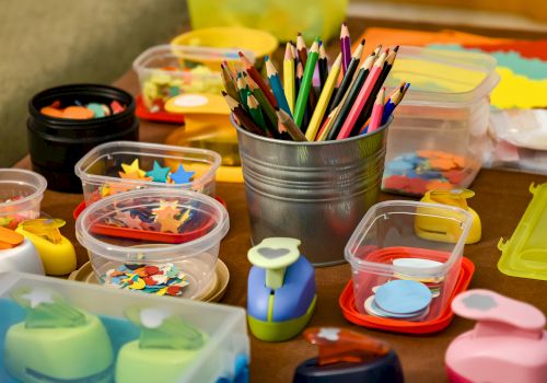 The image shows craft supplies, including colored pencils, various containers of cut-out shapes, and crafting tools on a table.
