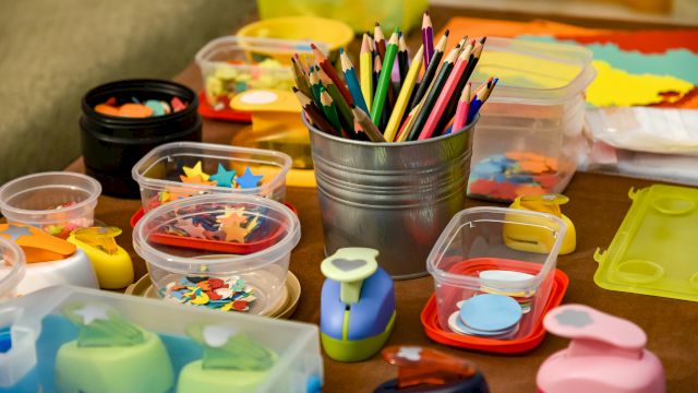 The image shows craft supplies, including colored pencils, various containers of cut-out shapes, and crafting tools on a table.