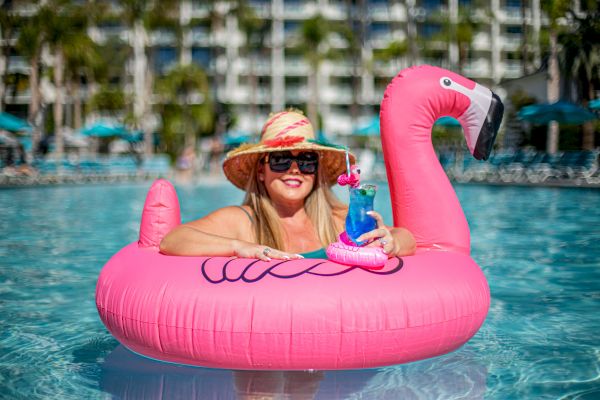 A person relaxes in a pool on a pink flamingo float, wearing a sunhat and sunglasses, holding a drink with an umbrella.
