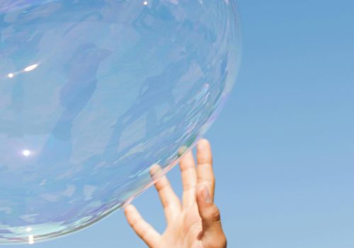 A hand reaching up towards a large, translucent bubble against a clear blue sky.