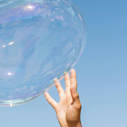 A hand reaching up towards a large, translucent bubble against a clear blue sky.