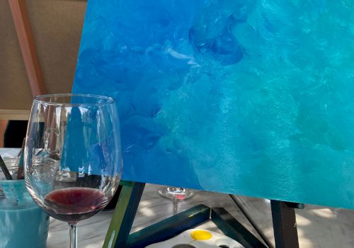 A painting setup features a canvas with blue tones on an easel, a paint palette with colors, and a half-full wine glass on the table.