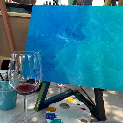 A painting setup features a canvas with blue tones on an easel, a paint palette with colors, and a half-full wine glass on the table.