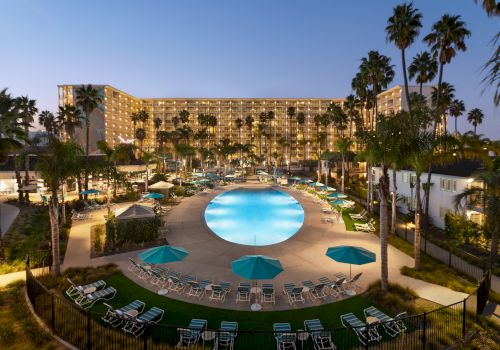 A large hotel with a glowing pool at dusk, surrounded by palm trees, loungers, and umbrellas, with the building lit in the background.