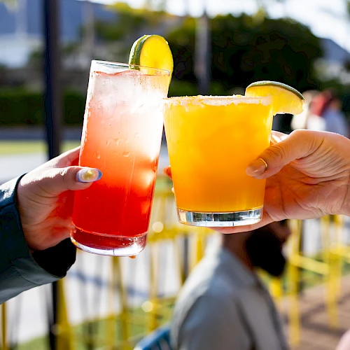 Two hands toasting with colorful cocktails, one red and one orange, garnished with lime and lemon slices, outdoors.