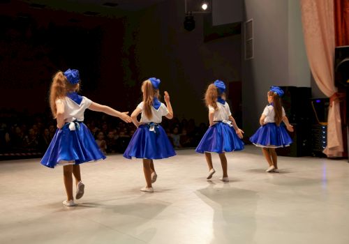Four children in blue and white costumes perform a dance on stage with an audience watching.