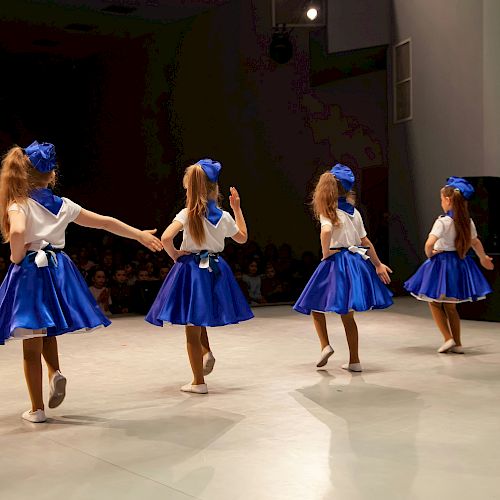 Four children in blue and white costumes perform a dance on stage with an audience watching.