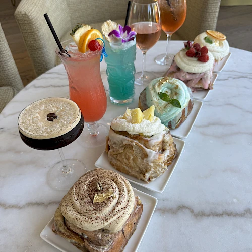 The image features a variety of colorful drinks and plates of pastries, including iced beverages with fruit garnishes and frosted cinnamon rolls.