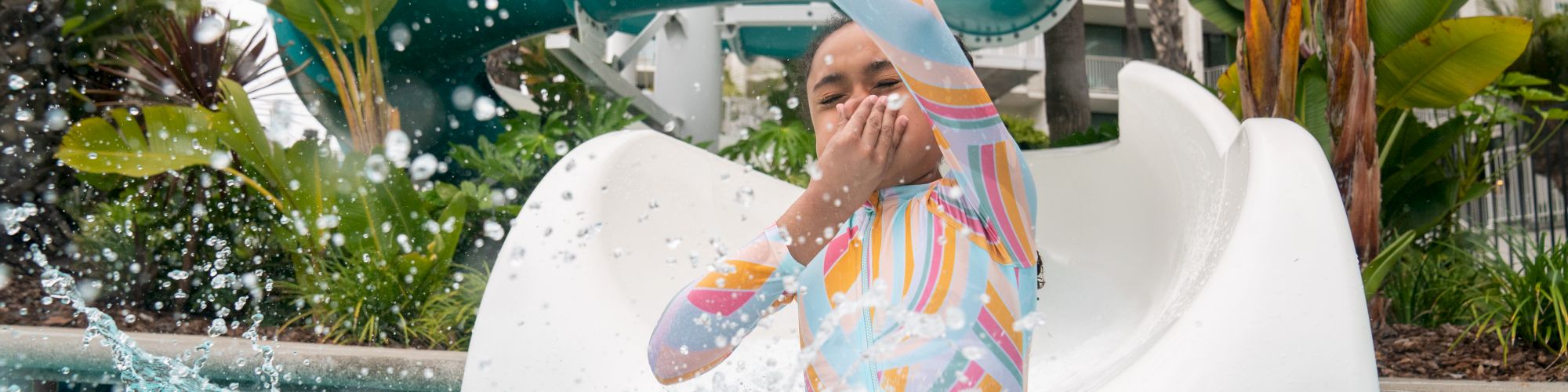 A person splashes into a pool at the bottom of a waterslide, surrounded by foliage and vibrant water activity.