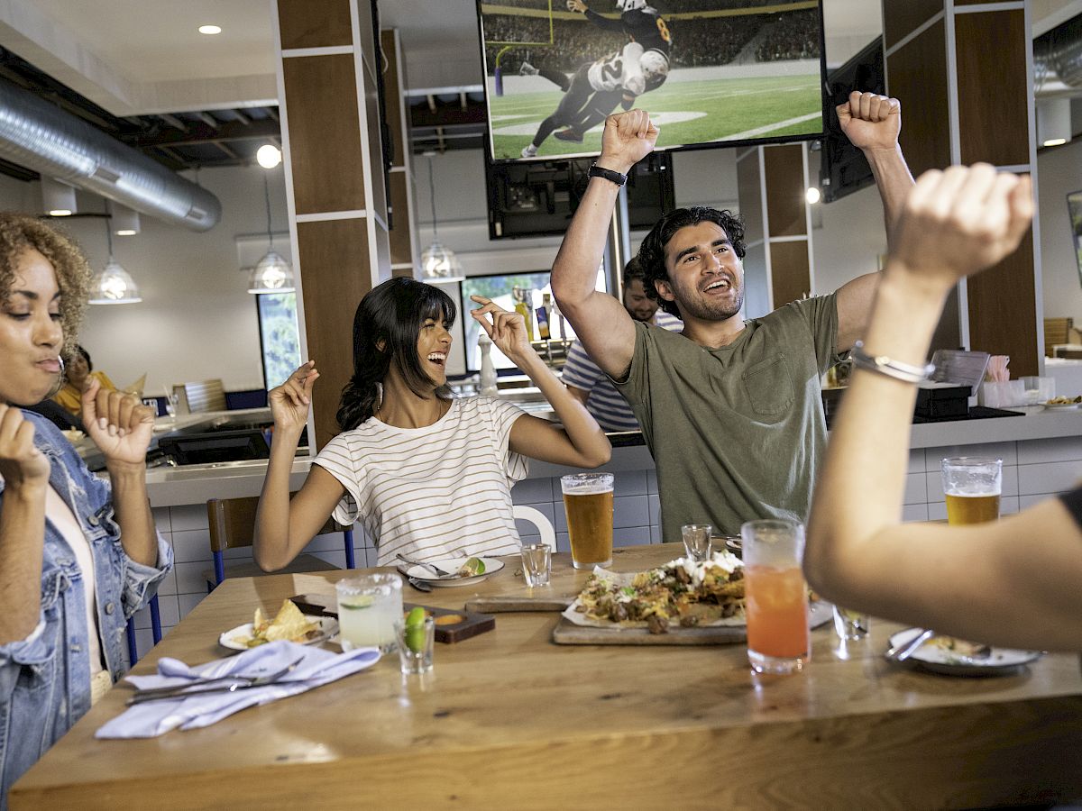 A group of people at a restaurant table, cheering and eating, with a football game on TV in the background.