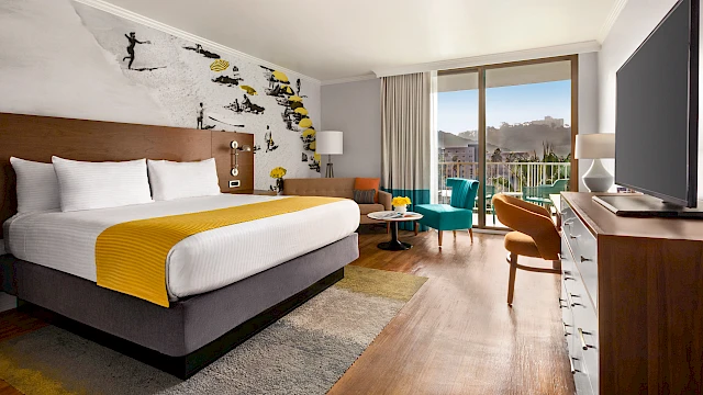 A hotel room features a large bed, snowy wall art, TV, sitting area, and balcony with daylight streaming in from glass doors.