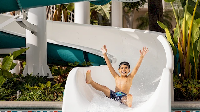 A child joyfully slides down a water slide into a pool. The slide is labeled "Twister." Tropical plants are in the background.