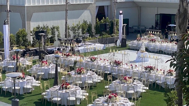 An outdoor event setup with round tables, white tablecloths, and floral centerpieces on a grassy area, next to a large open space.