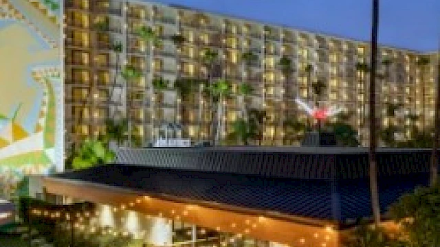 The image features a well-lit modern building with a rooftop bar area and a mural on the adjacent wall, surrounded by palm trees at night.