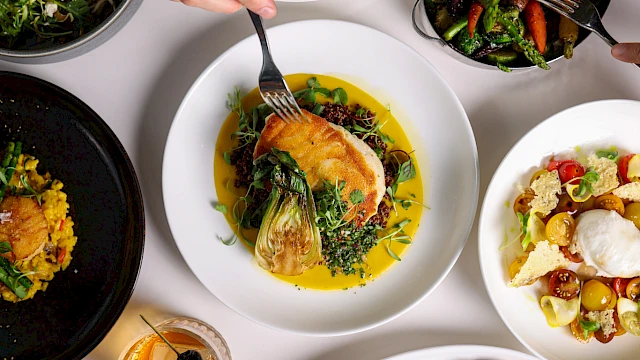 An assortment of dishes including meats, vegetables, and desserts, accompanied by drinks, on a table with a hand serving food from a plate.