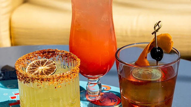 Three cocktails on a vibrant table: a yellow drink in a glass with a lime slice, a tall pink drink with fruit garnish, and a dark drink.