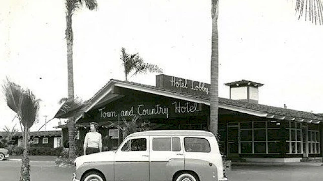 A vintage photo of a person posing by a classic car in front of the Town and Country Hotel, surrounded by palm trees.