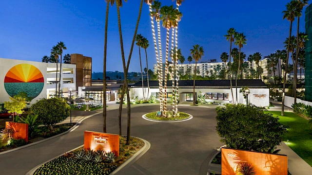 The image shows a hotel entrance at dusk with palm trees, modern landscaping, and a mural on a nearby building.
