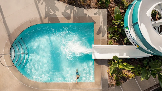 Aerial view of a person in a pool with a waterslide, surrounded by greenery and concrete.