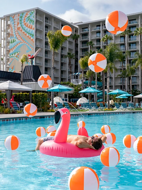 People relaxing on a sunny hotel pool deck with orange/white beach balls and an inflatable pink flamingo in the pool, palm trees in the background, and a multi-story resort. (140 characters, ending with a period.)