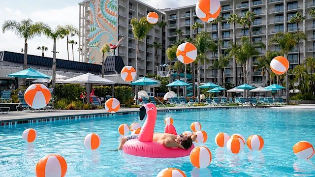 People relaxing on a sunny hotel pool deck with orange/white beach balls and an inflatable pink flamingo in the pool, palm trees in the background, and a multi-story resort. (140 characters, ending with a period.)